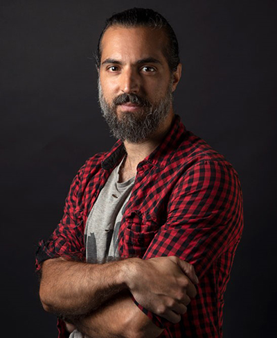 Studio portrait of Marco Libretti, young gentle with a salt and pepper beard, wearing a red and black checkered shirt, against a dark grey background. Photo by Candace Cosentino.