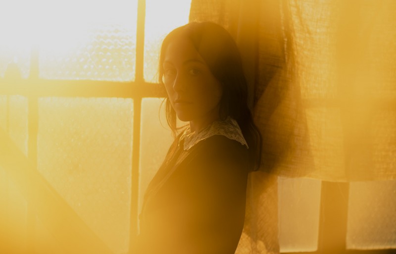 A portrait of Darby Milbrath, a young woman wearing a dark dress in front of an old paned warehouse window, with a warm glare of yellow and orange light. Photo by Norman Wong.