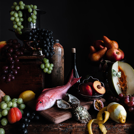 A picture of a table with pink fish, half cut peaches, cantaloupes, pineapples, oranges, bread, oysters along with green and black bunches of grapes