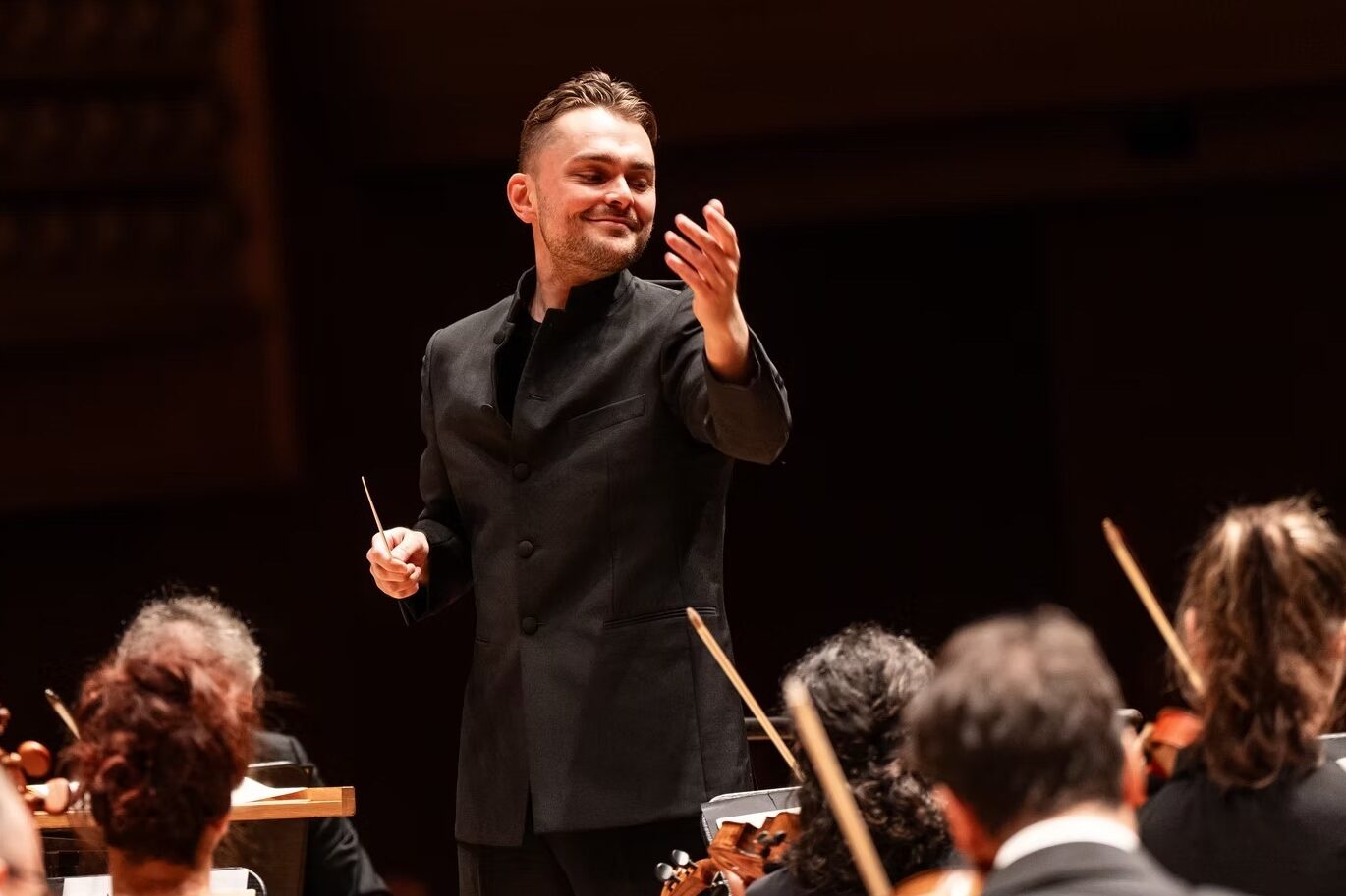 Jakob Lehmann faces an orchestra and smiles gently with one arm lifted to cue.